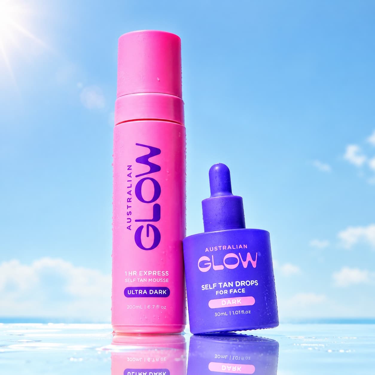 The It Couple — 1HR Express Ultra Dark mousse leaning together with Face Tan Drops against a summer sky